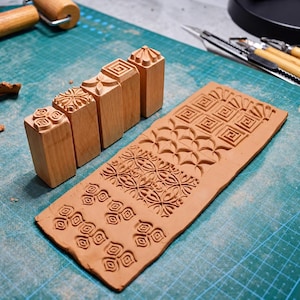 May include: Four light brown wooden stamps with square bases featuring carved geometric patterns, positioned next to a rectangular slab of light brown clay that displays the corresponding stamped impressions of these intricate designs.