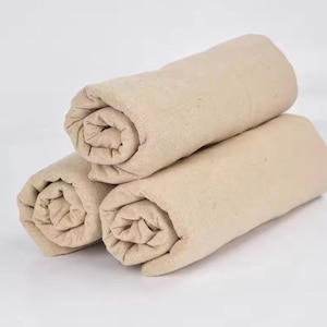 Puede incluir: Tres rollos de tela de color beige apilados juntos. Cada rollo está hecho de un tejido liso con una superficie visiblemente ligeramente texturizada. La tela es de color beige claro y parece estar enrollada firmemente en formas cilíndricas.