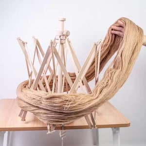 May include: A wooden yarn swift with tan yarn. The swift is open and ready to hold the yarn, which is being held by a hand. The swift is on a light brown table.