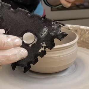 Op de afbeelding: Een zwart, plat, tandwielvormig aardewerk gereedschap met meerdere ingekeepte randen en een rond gat in het midden, geplaatst naast een lichtgekleurde, gladde, ronde kleikom op een pottenbakkersschijf, met resten van natte klei op het oppervlak.
