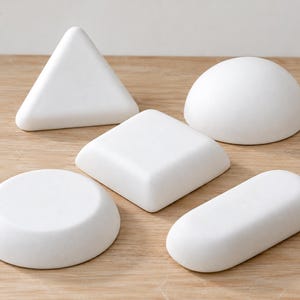 Op de afbeelding: Een verzameling van vijf massieve witte geometrische objecten, waaronder een driehoekig prisma, een halve bol, een vierkant blok met afgeronde randen, een ronde schijf en een langwerpige ovale vorm, allemaal met een gladde, matte witte afwerking.