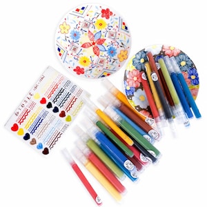 May include: A set of ceramic underglaze pens in various colors, a rectangular instruction sheet with heart and line patterns, and two white ceramic plates decorated with colorful floral and geometric designs in red, blue, and yellow.