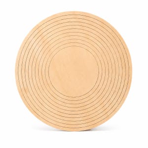 May include: A circular light-colored wooden board featuring a series of concentric engraved rings that radiate from the center toward the outer edge, creating a pattern of thin, dark lines across the smooth, flat surface of the wood.