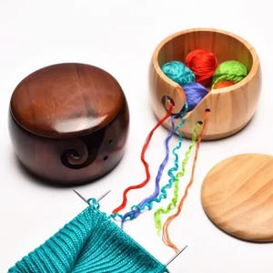 Puede incluir: Dos cuencos de hilo de madera, uno con tapa y diseño de espiral, el otro con ovillos de hilo de colores. Hilos de lana roja, azul, verde y naranja se extienden desde el segundo cuenco. Un proyecto de tejido turquesa está en primer plano.