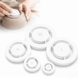 May include: A set of five white circular pottery centering tools in sizes XS, S, M, L, and XL. Each tool features a central disc connected to an outer ring by three spokes, with the size clearly embossed on the center of each white plastic piece.