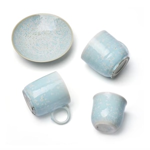 May include: A set of ceramic tableware featuring a light blue glaze with a white speckled pattern. The collection includes one round saucer and three mugs of varying sizes, all displaying a consistent mottled texture and smooth, rounded edges.