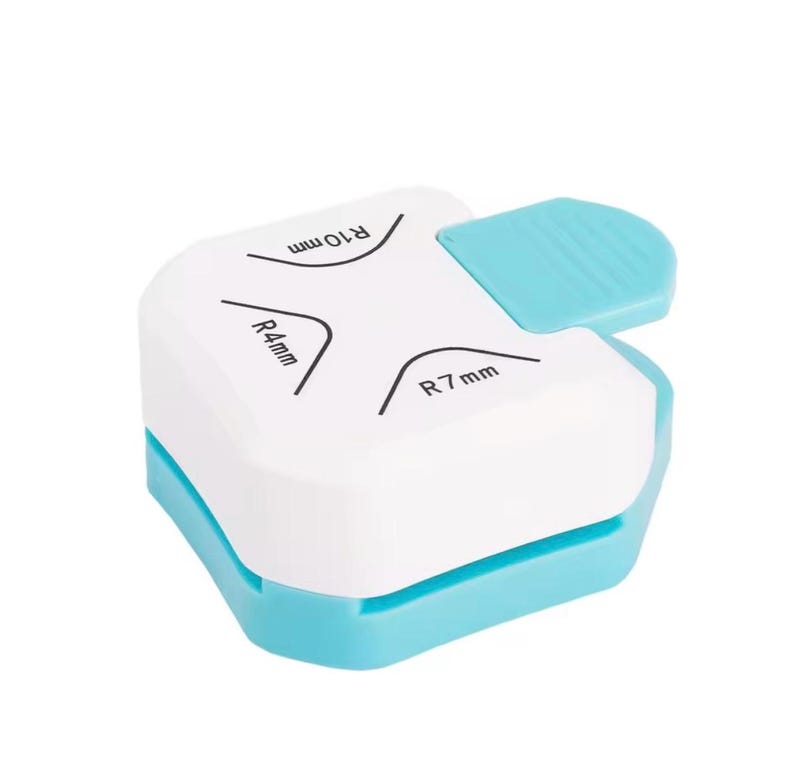 May include: A white and light blue corner rounder tool. The tool has a white top with black text indicating corner radius sizes of R4mm, R7mm, and R10mm. A light blue lever is on the top right. The base is light blue.