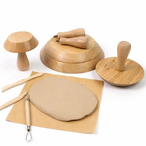 Puede incluir: Un conjunto de herramientas de cerámica de madera, que incluye una forma de hongo, un sello redondo y herramientas de modelado. Una losa de arcilla marrón descansa sobre papel marrón. Las herramientas son de madera clara.