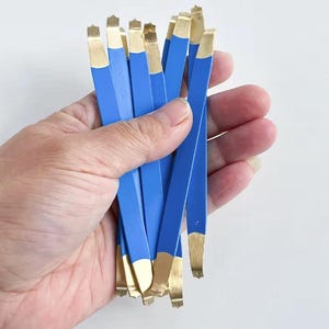 May include: A collection of multiple thin, flat metal strips featuring a bright blue center section and metallic gold-colored tips at both ends. These narrow, elongated tools have a smooth finish and are held together in a bundle.