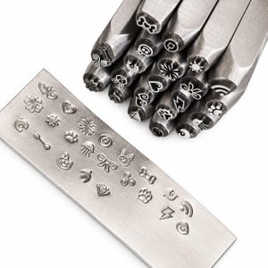 May include: A collection of silver-toned metal stamping tools with various engraved symbols on the ends, accompanied by a rectangular metal plate displaying the corresponding impressions of a heart, flower, sun, paw print, diamond, and lightning bolt.