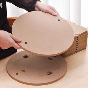 May include: A stack of circular light brown wooden boards with smooth surfaces. Each board features two small circular holes near the edges and a square cutout section in the centre. The material has a matte finish and a consistent, neutral tone.