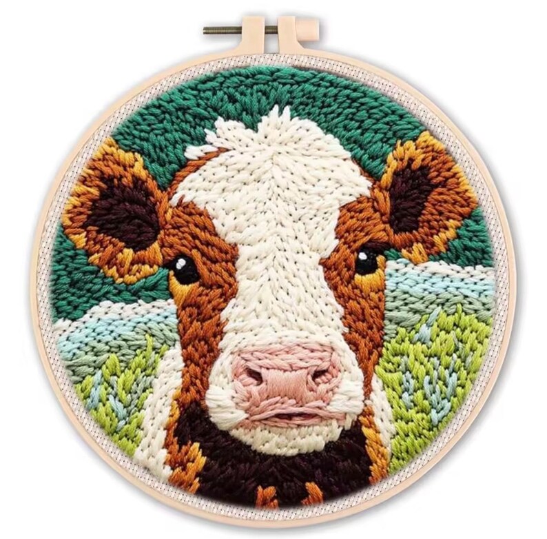 May include: A handcrafted embroidery hoop art piece featuring a detailed cow face. The cow has a white and brown face with a pink nose, set against a green and blue background. The embroidery is in a round wooden hoop.