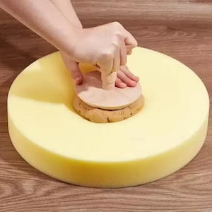 May include: A person using a wooden tool to shape clay on a large, round, yellow sponge. The tool has a circular base and a handle. The clay is a light brown color.