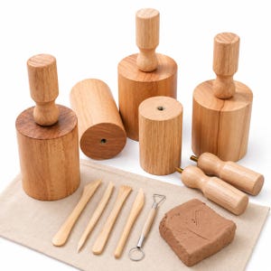 Puede incluir: Una colección de herramientas de alfarería de madera marrón claro que incluye varios sellos cilíndricos con mangos, cuatro palos de modelar de madera, una herramienta de lazo de alambre con mango de madera y un bloque rectangular de arcilla marrón sobre una tela beige.