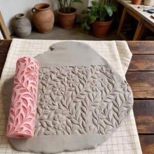 Könnte beinhalten: Eine zylindrische, rosafarbene Ton-Texturwalze mit eingraviertem Blumen- und Blattmuster, die auf einer flachen, grauen Tonplatte liegt. Die Walze zeigt ein sich wiederholendes Design aus kleinen Gänseblümchen und Blattreben, das in den Ton eingeprägt ist.