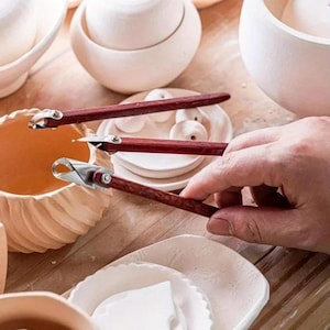 Könnte beinhalten: Nahaufnahme von Töpferwerkzeugen mit Holzgriffen und Metallklingen. Die Werkzeuge werden verwendet, um eine Keramikschale zu formen. Weitere Keramikteile, darunter Schalen und Teller, sind im Hintergrund zu sehen.