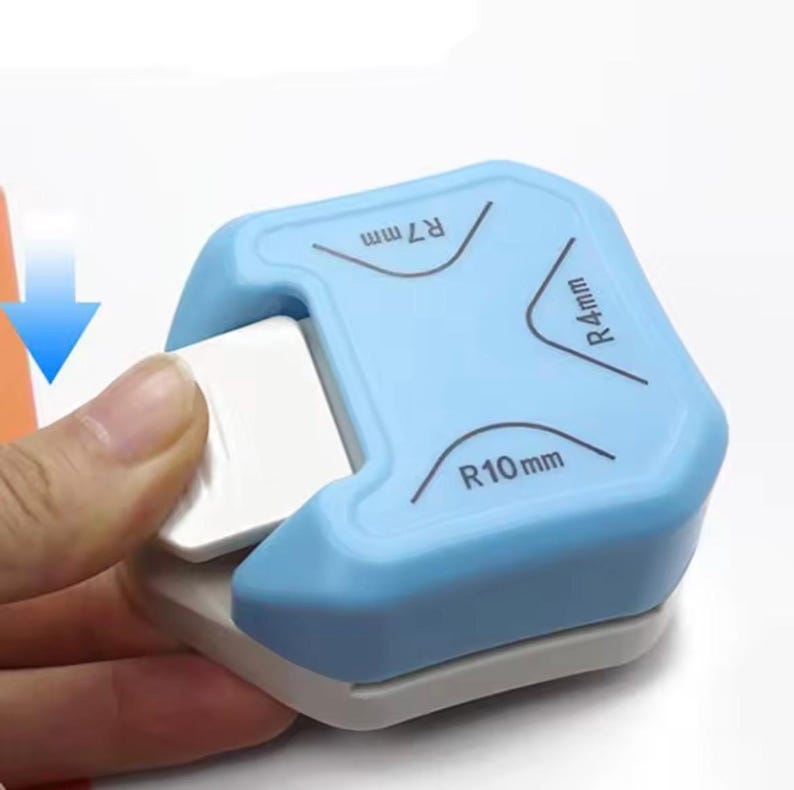 May include: A light blue and gray corner rounder tool. The tool has curved markings for R7mm, R4mm, and R10mm. A hand is shown using the tool to round the corner of a white piece of paper. A blue arrow points to the paper.