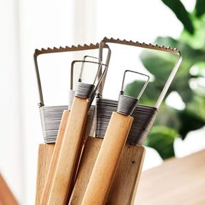May include: A set of pottery tools with wooden handles and metal blades. The blades have a serrated edge and are attached to the handles with coiled wire. The tools are displayed in a wooden holder.