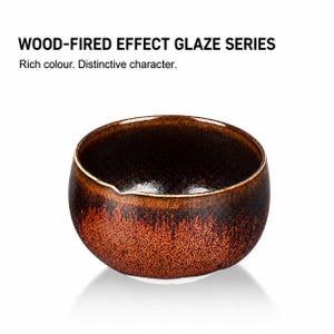 May include: A round ceramic bowl with a wood-fired effect glaze featuring a dark brown interior and a textured, mottled orange and black exterior. The text above reads: WOOD-FIRED EFFECT GLAZE SERIES. Rich colour. Distinctive character.