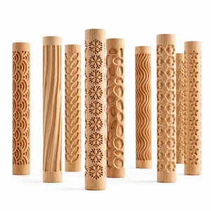 May include: A collection of nine cylindrical wooden rolling pins, each featuring a distinct carved pattern including snowflakes, flowers, stars, waves, leaves, and geometric shapes, arranged upright in a group against a white background.