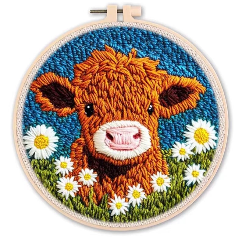 May include: Embroidered artwork featuring a brown cow with a pink nose and dark brown ears, surrounded by white daisies and green grass. The background is a textured blue. The artwork is set in a round wooden embroidery hoop.