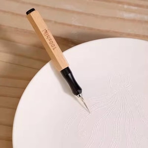 May include: A craft tool with a light wood hexagonal handle featuring the text 100shiki, a black metal grip, and a sharp metal needle tip, positioned on a white ceramic plate with a delicate embossed floral pattern on its surface.