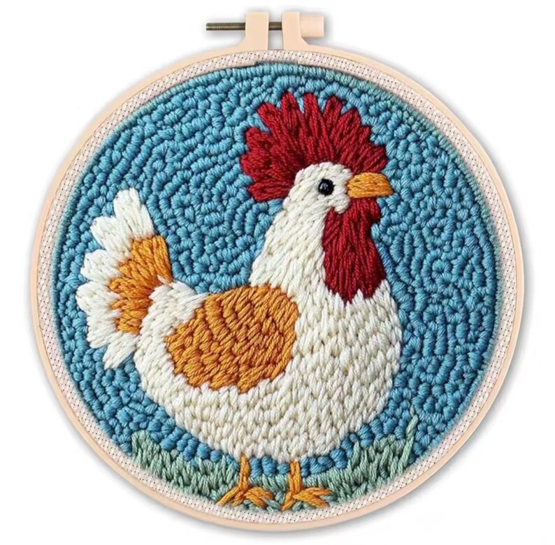 May include: A colorful rooster embroidery hoop art. The rooster features a red comb and wattle, white body, orange and white tail feathers, and orange accents. The background is blue, and the rooster stands on green grass.