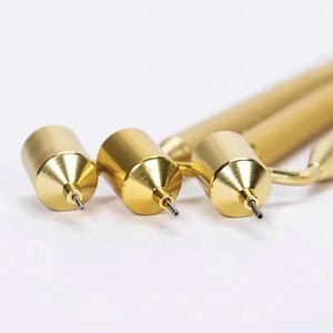 May include: Three gold-colored mechanical pencils with cylindrical bodies and conical tips. Each pencil features a thin, exposed lead point extending from the center of the metallic tip, showcasing a polished, reflective finish on the metal surface.