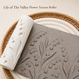 Könnte beinhalten: Ein weißer Texturroller mit Maiglöckchen neben einer grauen Oberfläche mit demselben Blumenmuster. Der Roller und die Oberfläche befinden sich auf einem Weidenkorb. Der Text "Lily of The Valley Flower Texture Roller" steht oben.