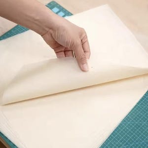 Puede incluir: Una pila de hojas de papel rectangulares, translúcidas y de color blanco hueso. La hoja superior está parcialmente levantada, revelando su textura fina y suave y su color crema claro. El papel está colocado sobre una base de corte verde con cuadrícula.