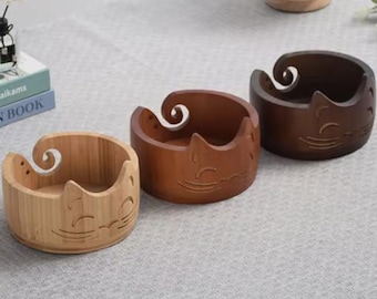 May include: Three wooden yarn bowls in varying shades of brown. Each bowl features a cat face design carved into the side and a swirl-shaped yarn guide. The bowls are made of bamboo and wood.