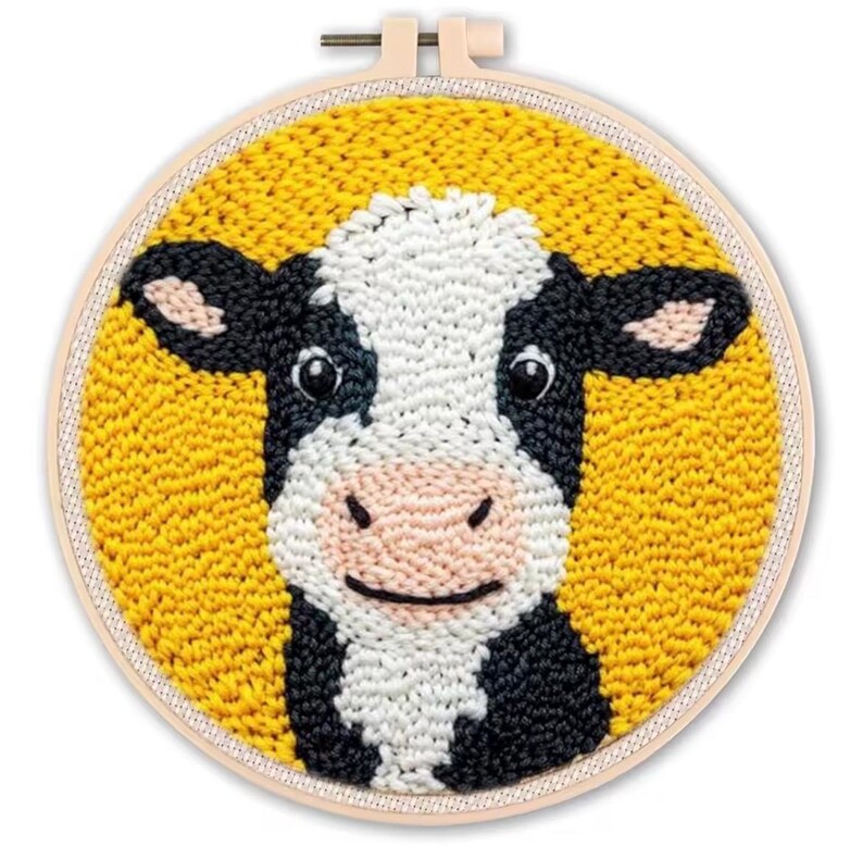 May include: A punch needle embroidery hoop featuring a smiling cow face. The cow has black and white markings, a pink nose, and black eyes. The background is yellow, and the hoop is made of wood.