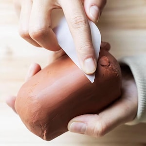 May include: A block of terracotta-colored modeling clay being smoothed with a flexible, white, triangular plastic rib tool. The clay has a dense, matte texture, and the tool is held against the surface to create a flat, even finish on the material.