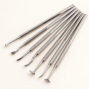 May include: A set of seven silver-colored metal sculpting tools arranged in a fan pattern. Each tool features a long, cylindrical handle with a textured, knurled grip section and a unique, shaped metal tip designed for precision work and detailing.