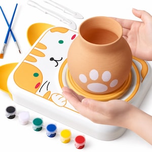 May include: A pottery wheel kit featuring a white base with a yellow cat illustration, a terracotta clay pot with a white paw print, two blue paintbrushes, two white sculpting tools, and a strip of six small paint pots in various colors.