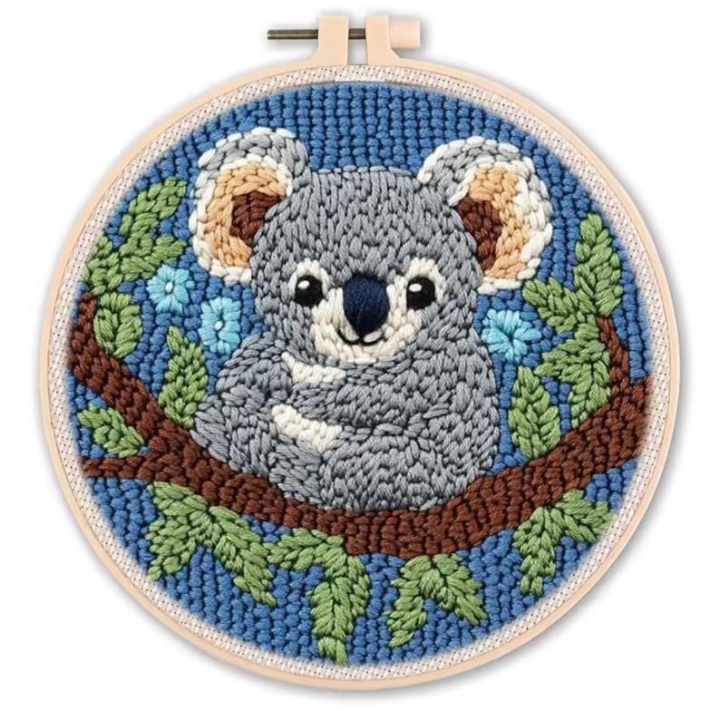 May include: A round embroidery hoop featuring a gray koala bear sitting on a brown branch, surrounded by green leaves and blue flowers. The background is a textured blue fabric. The koala has a white belly and tan ears.
