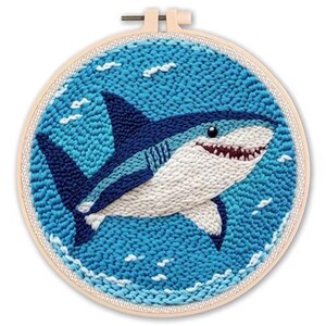 May include: A hand-stitched embroidery hoop featuring a shark design. The shark is depicted in shades of blue and white, with a red-lined mouth and a friendly expression. The background is a textured blue, with white accents.