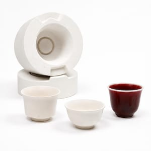 May include: A white plaster ceramic mold consisting of two stacked cylindrical parts, accompanied by two small matte white ceramic cups and one small glossy deep red ceramic cup, all featuring a simple rounded shape and a small base.