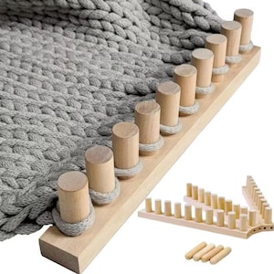 Puede incluir: Un telar de punto de madera con clavijas cilíndricas y una manta de punto gris. El telar tiene un acabado de madera clara y está diseñado para crear artículos de punto. También se muestran piezas de telar adicionales.