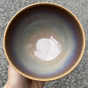 May include: A ceramic bowl with a lustrous, iridescent finish. The interior transitions from a light beige to a deep purple-blue at the rim. The rim has a golden-brown color.