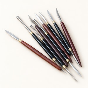 May include: A set of eight double-ended sculpting tools featuring polished metal tips and handles made of dark brown or black wood, connected by small brass ferrules. The tools include various shapes such as pointed blades, rounded ends, and ball tips.