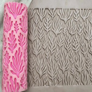 May include: A cylindrical pink silicone rolling pin featuring a raised, repeating seaweed or coral-like botanical pattern. Next to the roller is a flat, grey slab of clay that has been impressed with the same intricate, organic leaf-like design.