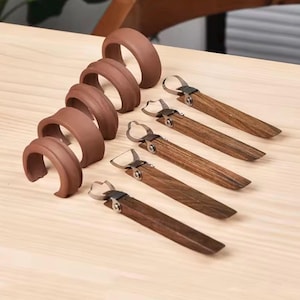 May include: A set of five matte brown C-shaped silicone napkin rings arranged in a vertical row, paired with five matching wooden-handled metal tools featuring a small, rounded wire loop at the end of each handle for securing napkins.