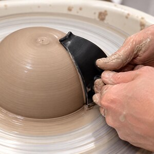 Könnte beinhalten: Ein schwarzes, gebogenes Töpferwerkzeug, das gegen eine nasse, hellbraune Tonschale auf einer rotierenden Töpferscheibe gehalten wird. Das Werkzeug verfügt über eine glatte, ergonomische Kante zum Formen und Glätten der äußeren Tonoberfläche.
