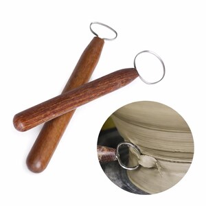 May include: Two pottery loop tools with smooth, dark brown wooden handles and thin, silver-colored metal wire loops at the ends. A circular inset shows one of the tools being used to trim excess gray clay from a piece on a pottery wheel.