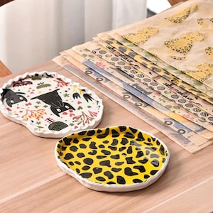May include: Two decorative ceramic trays with distinctive designs. One tray has a yellow leopard print, the other a whimsical pattern with black animal figures and botanical elements. A stack of patterned paper sheets is in the background.