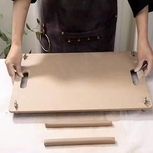 May include: A rectangular light brown wooden slab with two oval cut-out handles on the sides and four metal wing nuts at the corners. Two separate cylindrical wooden rods are positioned horizontally in front of the main rectangular board.