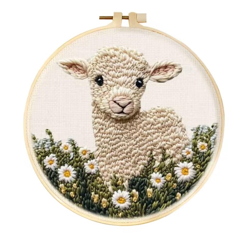 May include: Embroidered artwork of a lamb in a field of daisies, set in a wooden embroidery hoop. The lamb is beige with black eyes and a pink nose. The daisies have white petals and yellow centers.