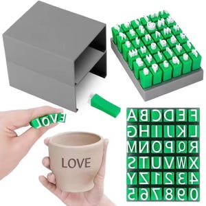 Puede incluir: Un juego de sellos del alfabeto en verde y blanco con una caja de almacenamiento gris y una bandeja organizadora gris. Los sellos incluyen letras y números individuales. Se muestra una taza de cerámica beige con la palabra LOVE grabada.