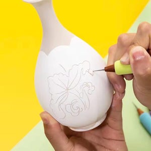 May include: A white ceramic vase with a narrow neck and a rounded body featuring a delicate, hand-drawn floral outline etched into the surface. A light green tool with a fine metal tip is being used to trace the intricate petal and leaf design.